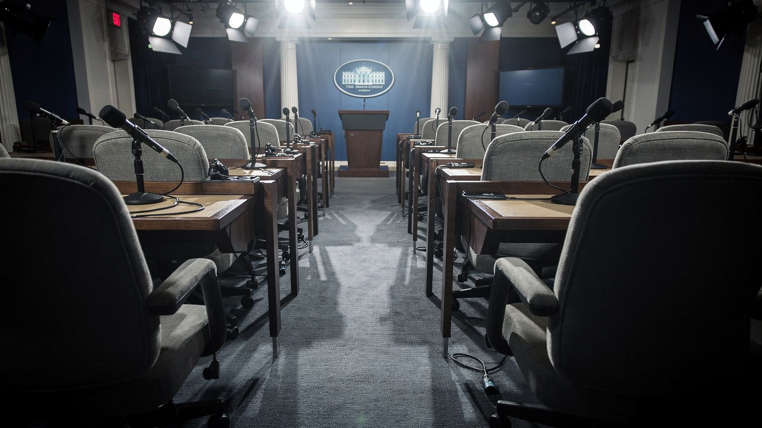 Rows of empty padded press briefing room seats in the White House Brady Press Briefing Room, each chair bare with no occupant, harsh overhead spotlights raking across the vacant desks and unused micro