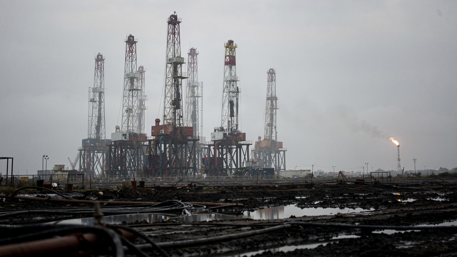 A cluster of oil drilling rigs stands silhouetted against a hazy, overcast Venezuelan sky, their steel latticework and pipes catching a cold, flat light, with a distant flare burning low and orange ne