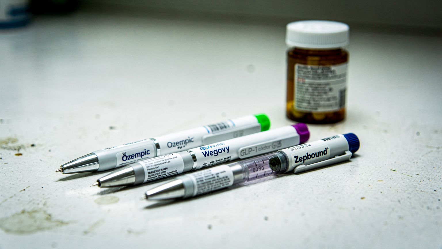 Three GLP-1 injector pens — Ozempic, Wegovy, Zepbound — lined up on a clinical countertop under harsh fluorescent light, their sleek bodies reflecting cool white glare, a prescription bottle blurred i