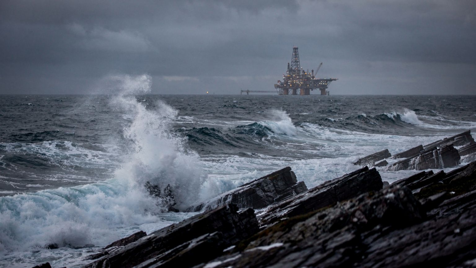 A windswept Falkland Islands coastline at dusk, a distant offshore oil platform silhouetted against a steel-gray sea, low-angle wide shot emphasizing the stark, exposed horizon, choppy waves crashing 
