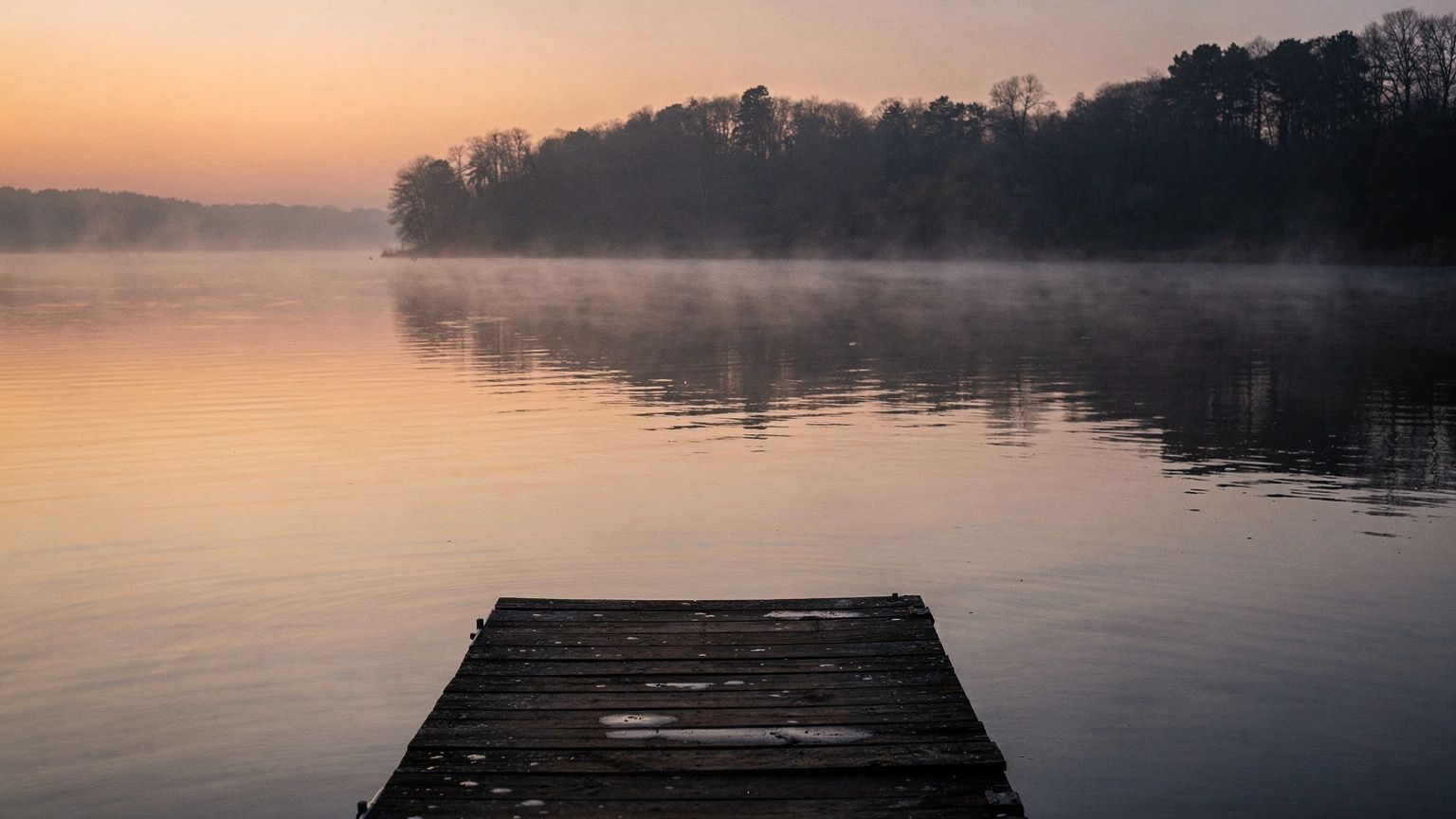 A still, somber wide shot of a warm freshwater lake at dusk, its glassy surface reflecting muted amber light, a lone wooden dock extending into the water, and a faint mist hanging over the shoreline, 