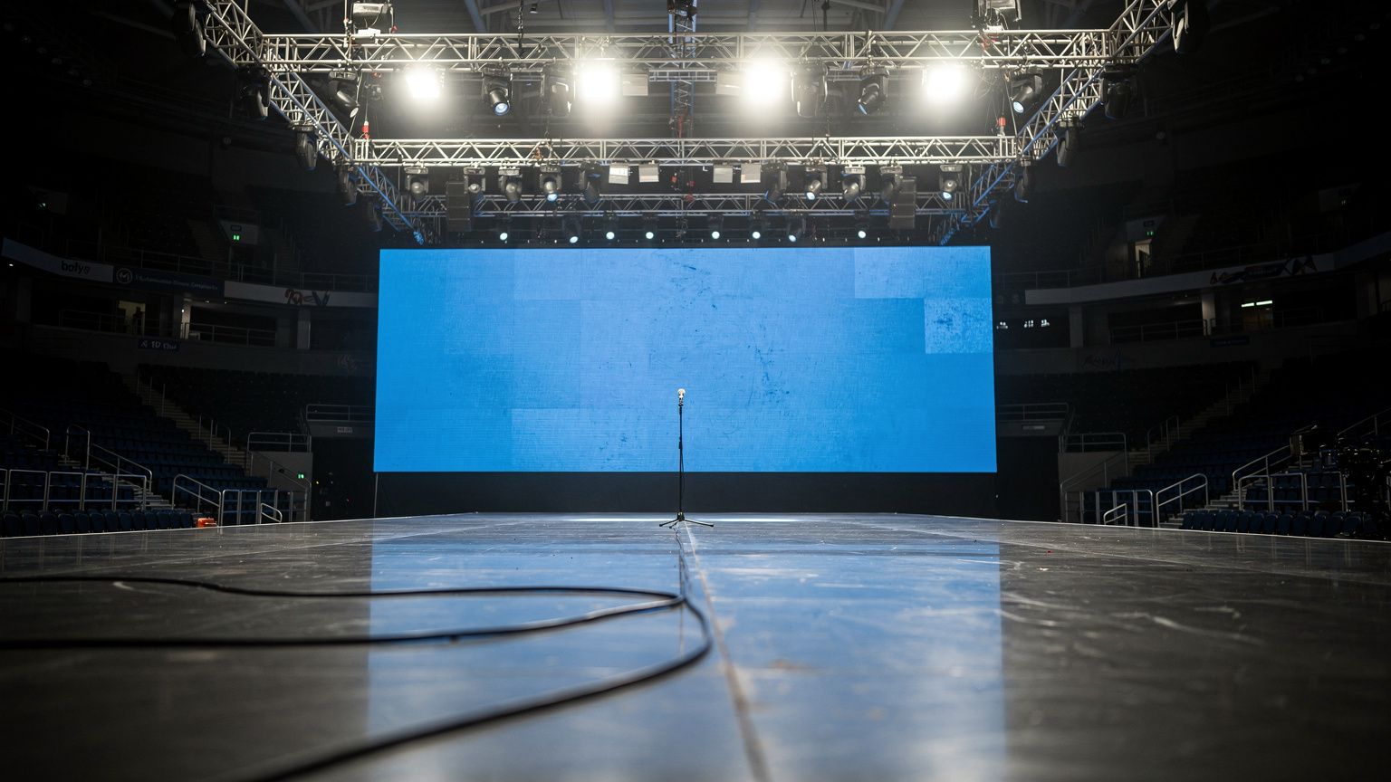The Eurovision Song Contest stage stands empty under harsh white work lights, its LED backdrop glowing a muted, contested blue, with a single microphone cable trailing across the polished floor, the v