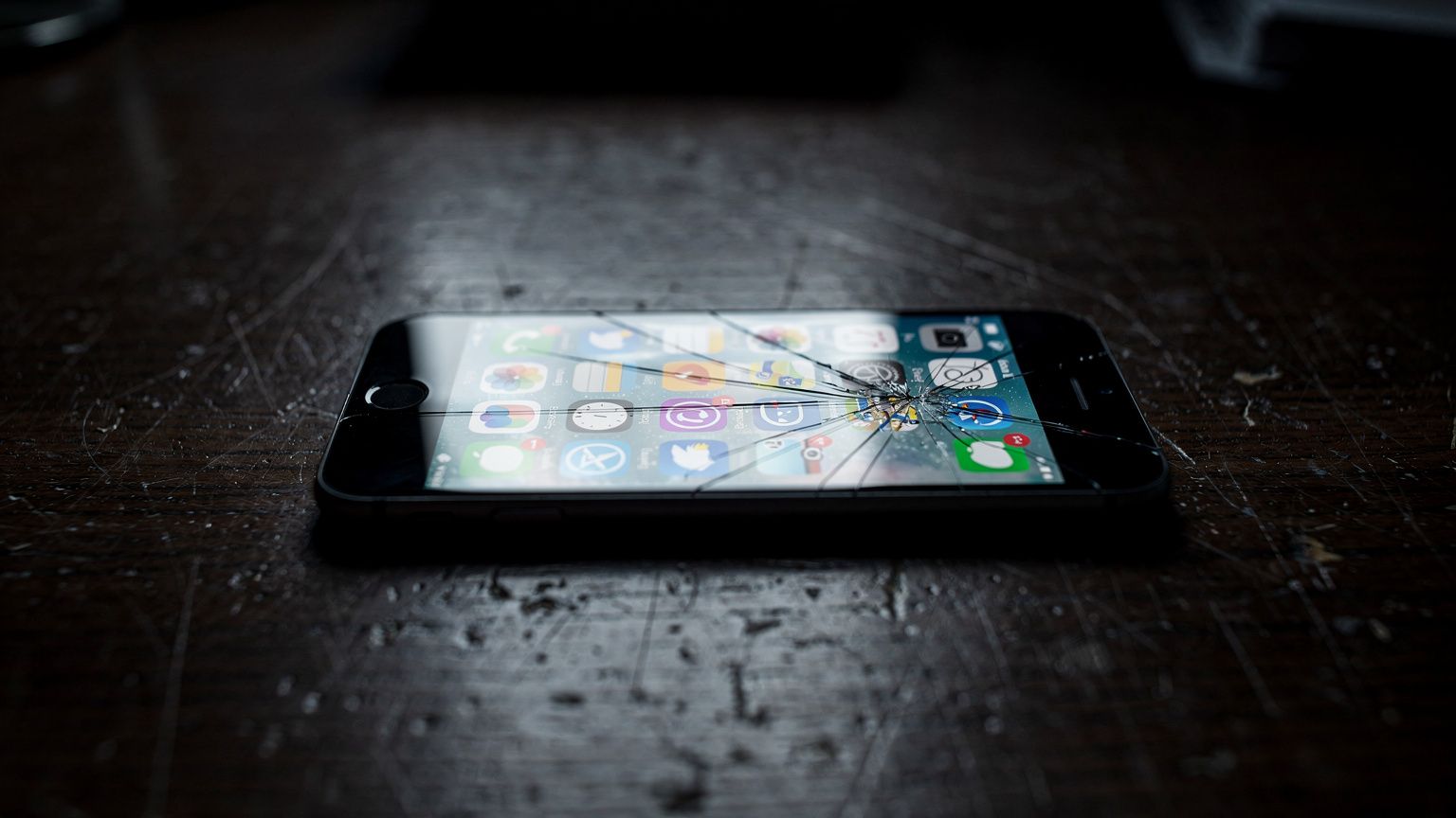 A smartphone lies face-up on a dark wooden desk, its screen a bright grid of social media app icons casting cold light across the surface, a single crack running diagonally across the glass, no people