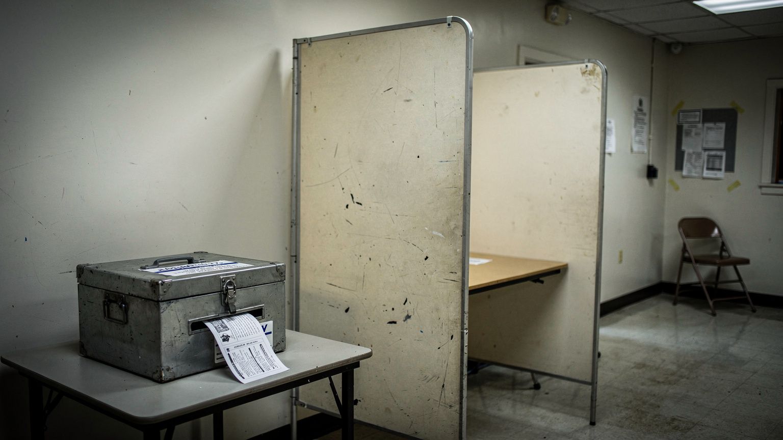 A Minnesota polling-place voting booth photographed empty and unattended, a single paper ballot resting half-inserted in the slot of a locked ballot box beside it, harsh overhead fluorescent light rak