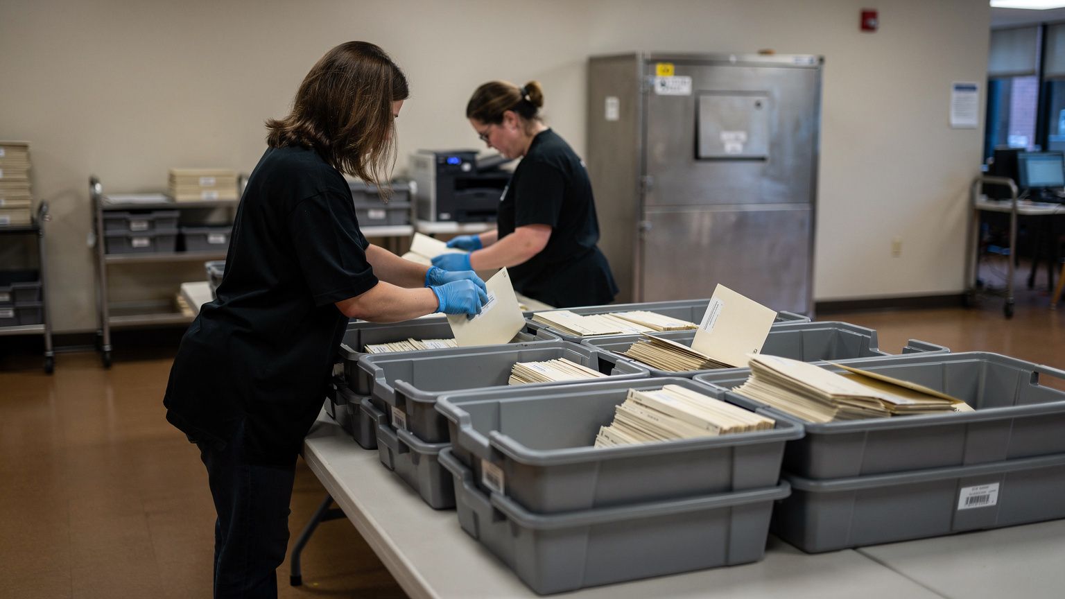 Two election workers in blue nitrile gloves, seen from behind and the side at a distance, sort and slit open off-white mail-in ballot envelopes from large muted gray plastic trays stacked on a long la