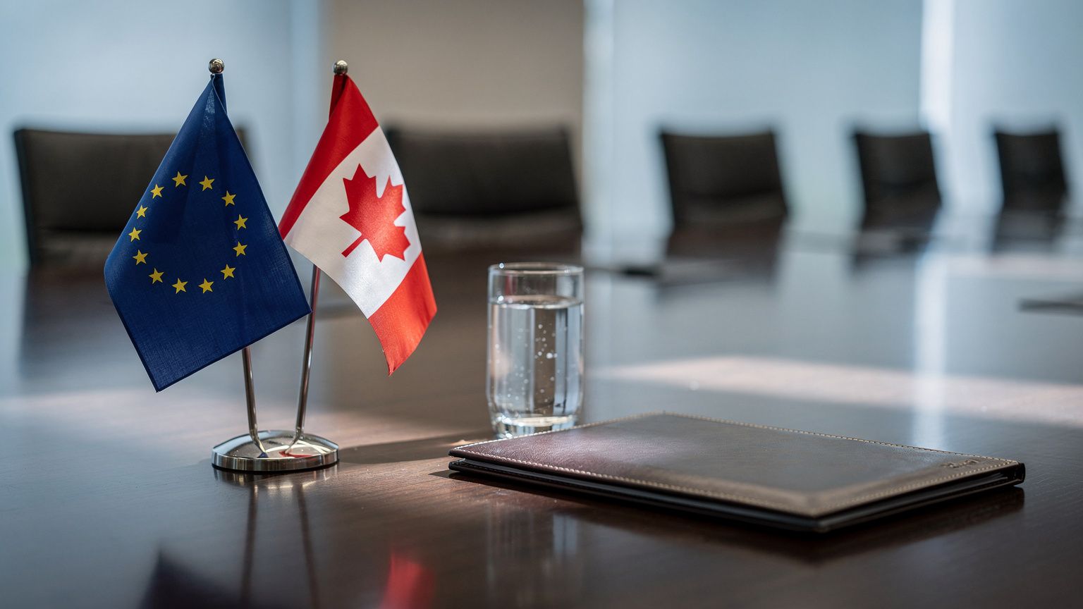 The European Union flag and the Canadian flag standing side by side in small tabletop stands on a polished summit conference table, an untouched glass of water and a closed leather folder beside them,
