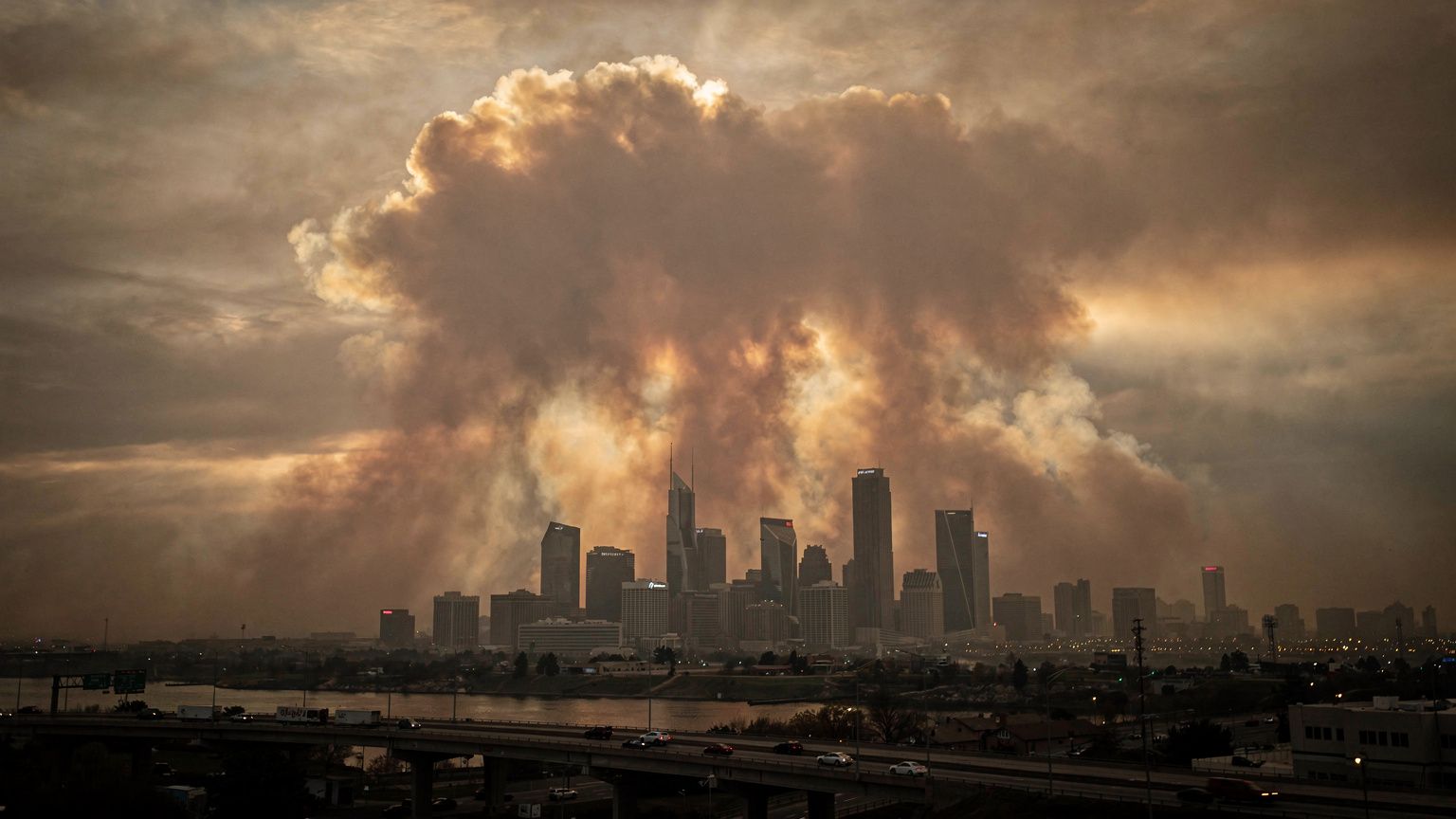 A vast wildfire smoke plume, thick and ochre, billows over a distant city skyline at dusk, the low sun casting a somber amber glow through the haze, wide-angle composition emphasizing the scale of the
