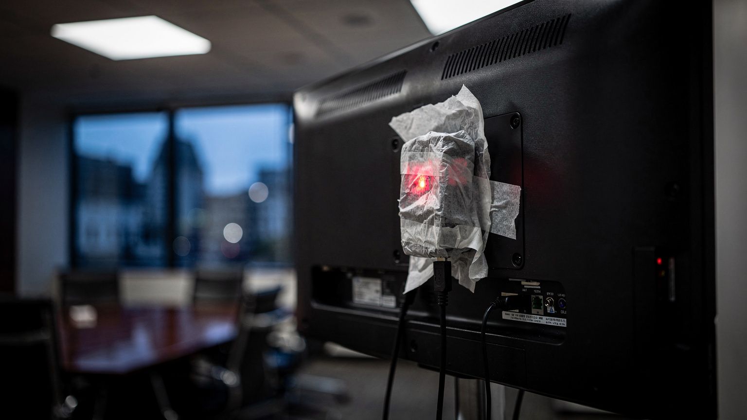 A small tissue-wrapped recording device taped over and plugged into the USB port on the back of a wall-mounted television in a dim, empty executive meeting room in San Francisco, its tiny indicator li