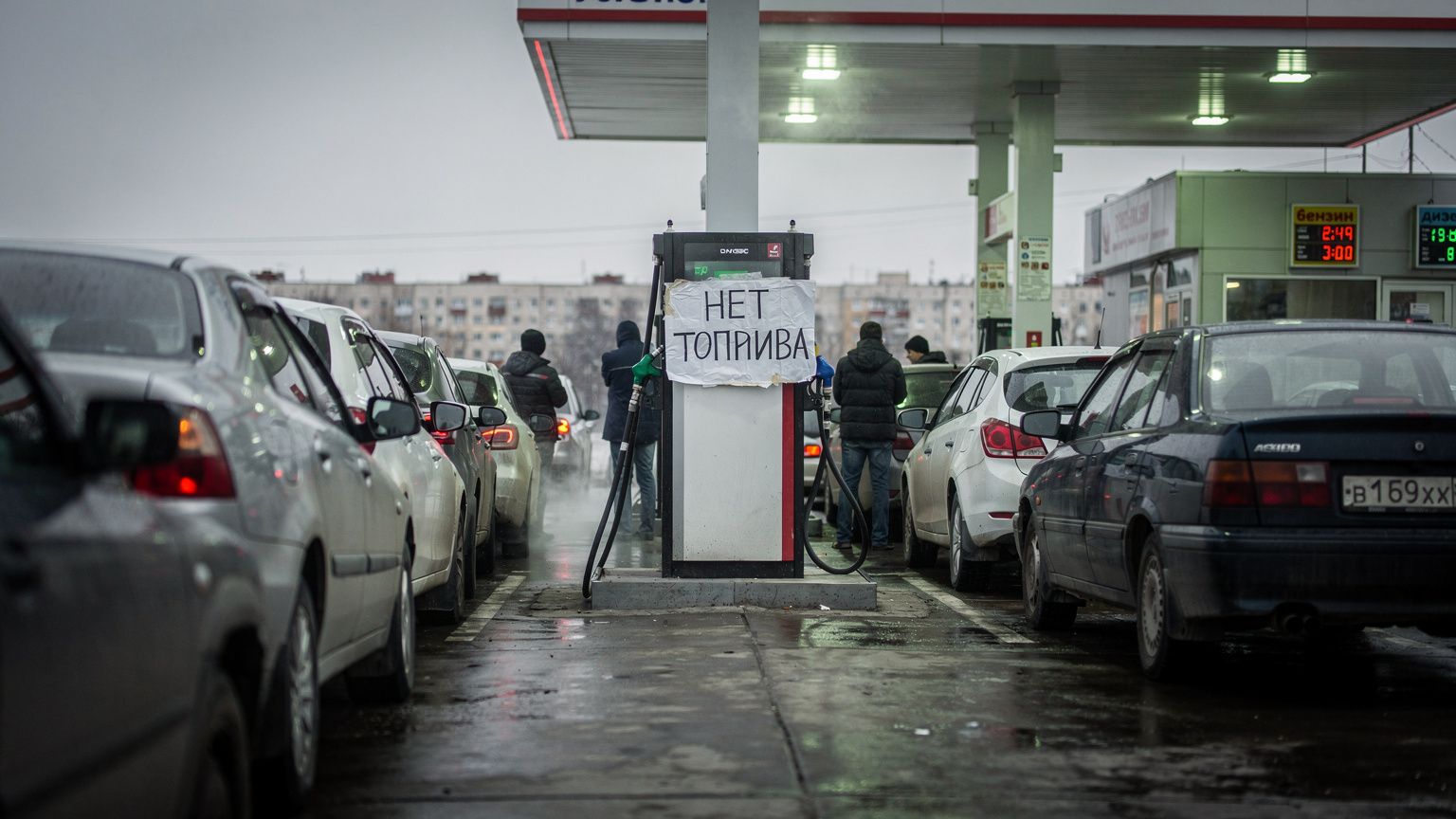 A long queue of parked cars at a Russian gas station under overcast skies, drivers standing beside their vehicles staring at a fuel pump with a handwritten 'no fuel' in Russian sign taped to it, the s