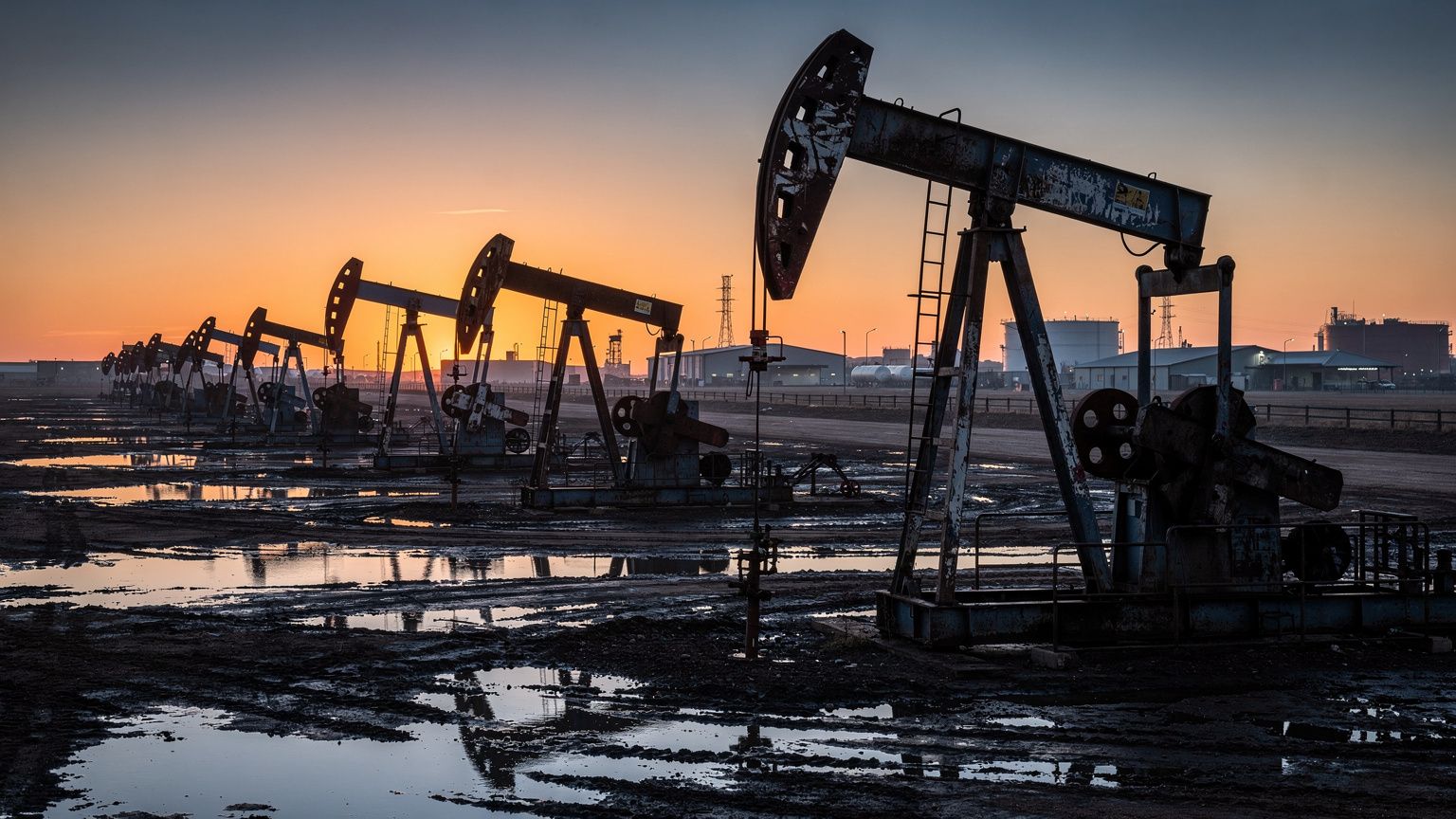A wide horizontal view of a sprawling oil field at dusk, rows of pump jacks silhouetted against a low orange sky, the nearest jack in sharp focus with its counterweight mid-stroke, wet ground reflecti