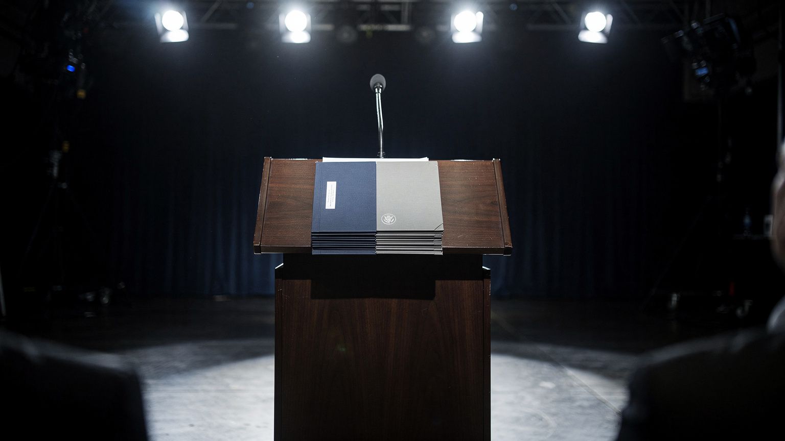A wide shot of a polished wooden lectern on a dark stage, empty and spotlit, with a single microphone and a stack of unopened briefing folders, the US Treasury seal faintly visible on the folders, har