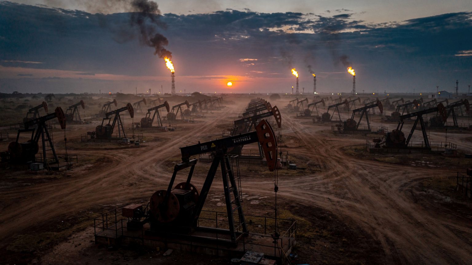 Aerial view of a sprawling Venezuelan oil field at dusk, rows of pumpjacks silhouetted against a low amber sun, one rig in sharp focus with its counterweight mid-stroke, distant flare stacks burning o