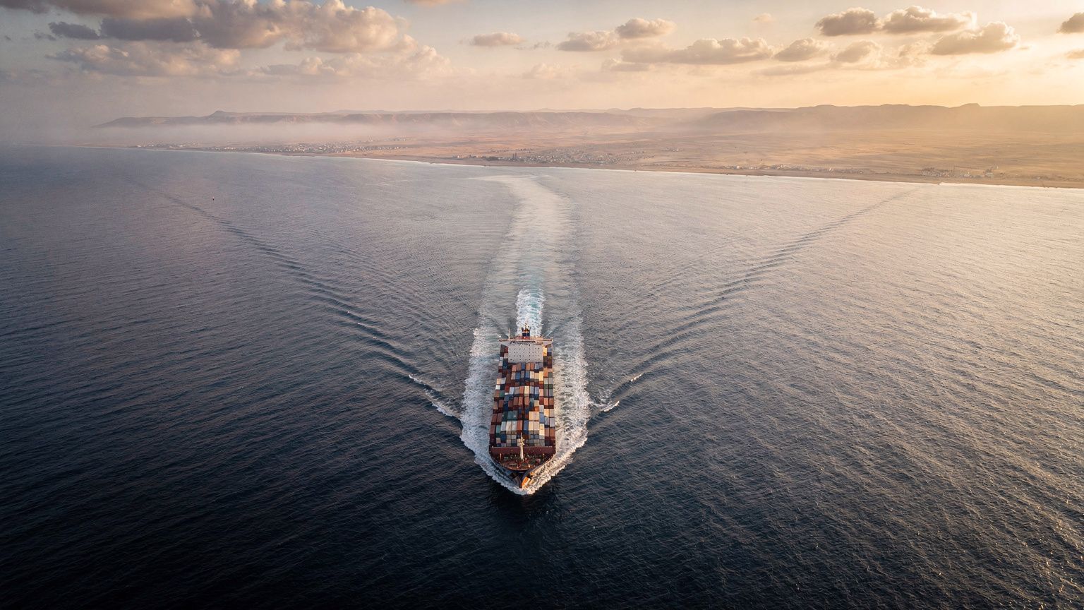 A wide aerial view of the Red Sea shipping lane off Yemen's coast at dawn, a lone container ship cutting a white wake through dark water, hazy desert coastline on the horizon, urgent low golden light 
